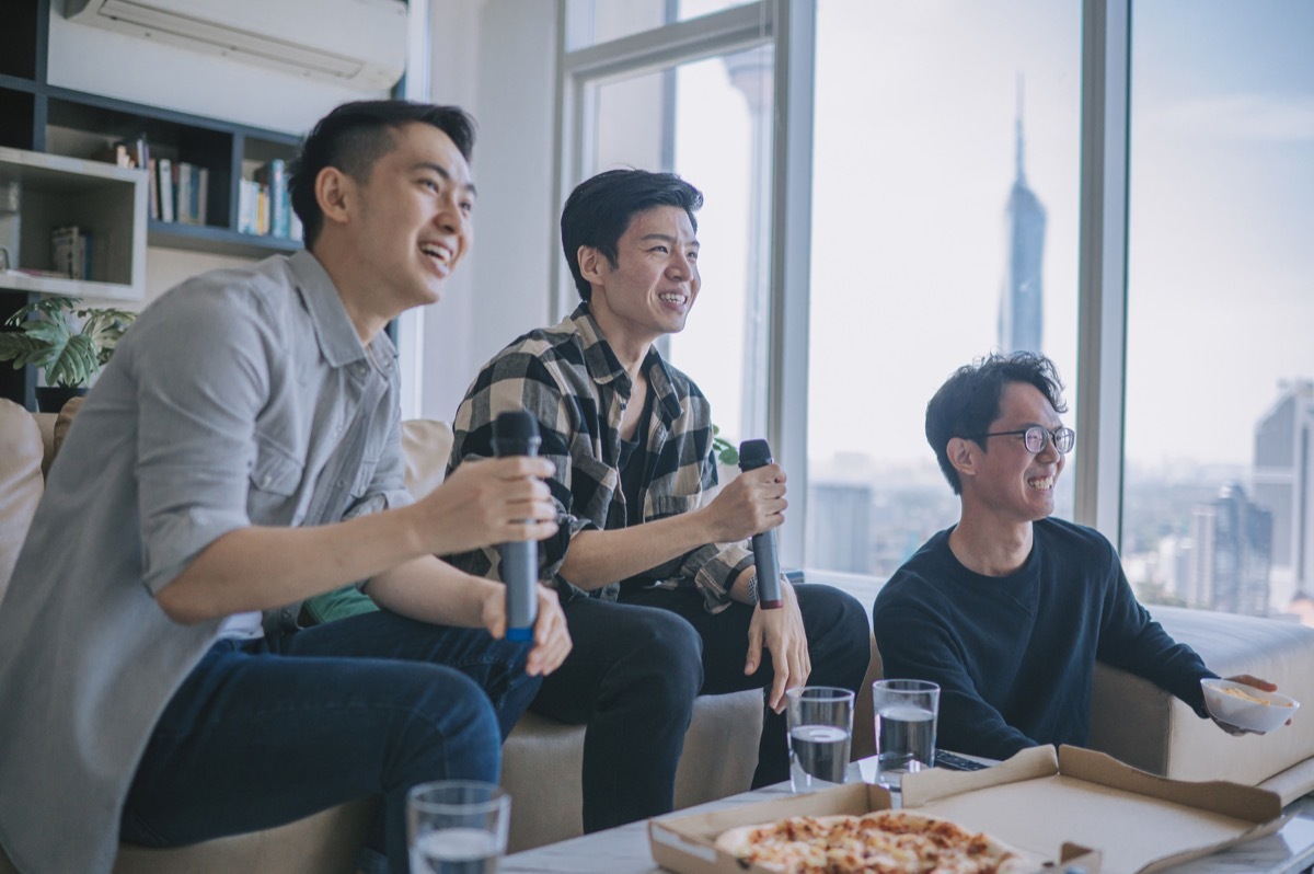 male friends enjoying karaoke session at home during weekend eating pizza