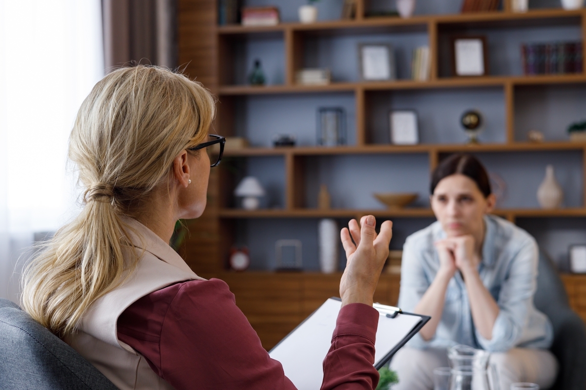 Over shoulder view of female psychologist sitting in armchair, talking with upset woman patient.