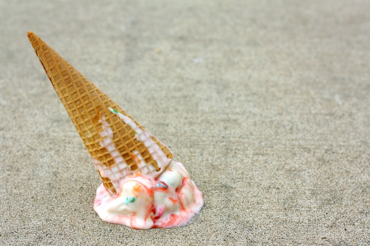 A rainbow colored ice cream cone has dropped upside down on the sidewalk on a summer day