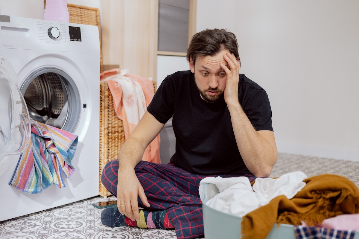 Man sits on the laundry room floor with the washing machine open, looking confused and overwhelmed.