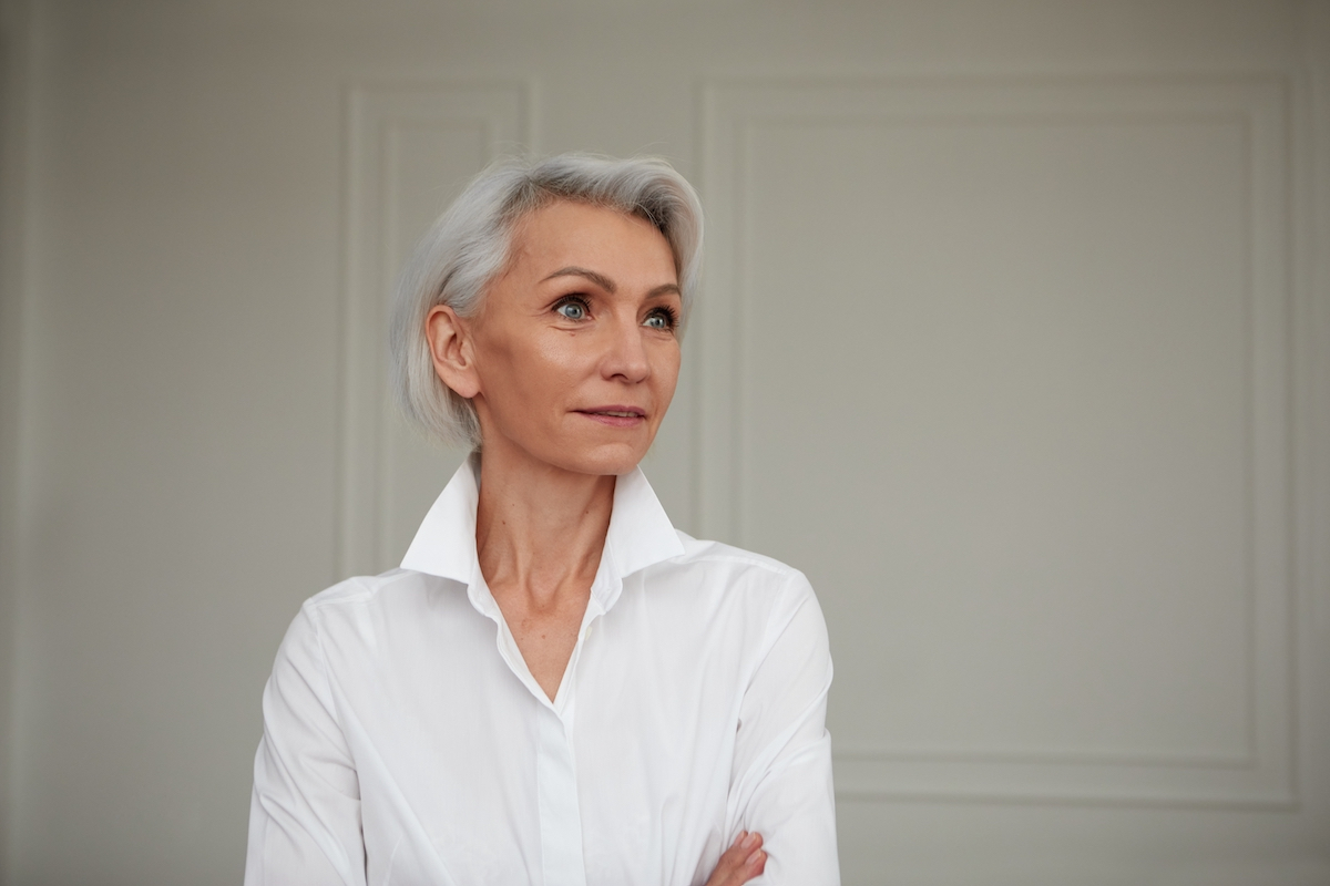 Portrait of an adult elderly self-confident woman with white hair looking awey into distance in white shirt hands crossed background wall