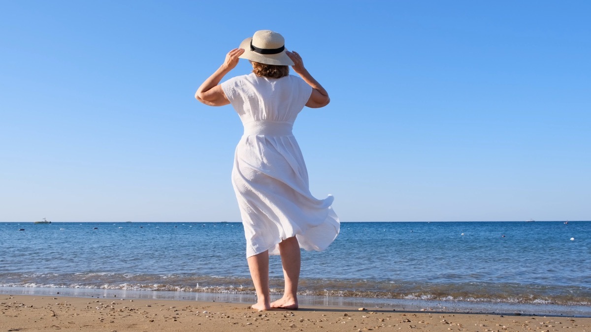 back view on a mature woman in a straw hat and white dress walks along the blue sea coast on a sunny summer day, enjoying freedom and relaxation. The concept of a typical life of pensioners.