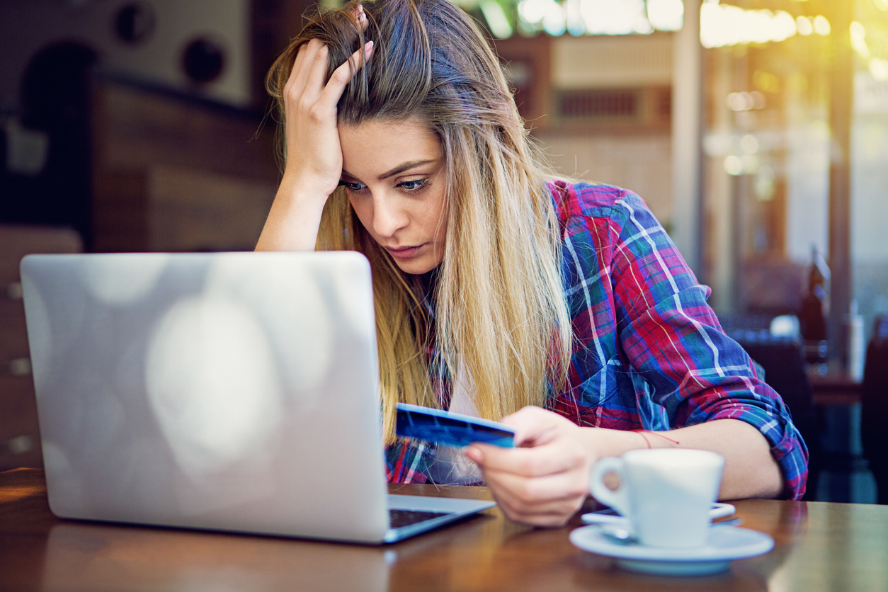 A young woman sitting at her laptop while holding her credit card with a stressed look on her face