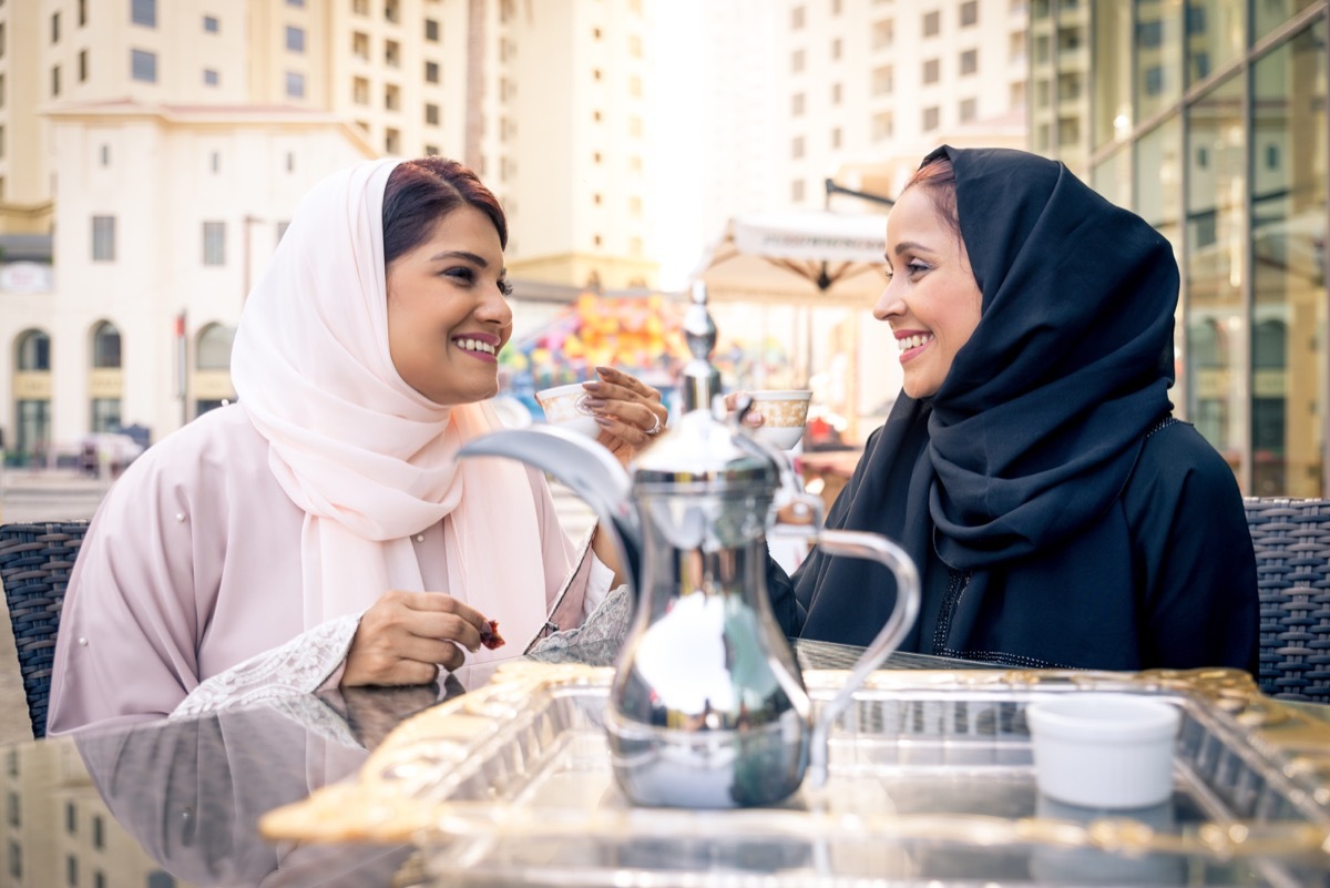 Two women wearing hijabs—one pink and one black—share tea outside