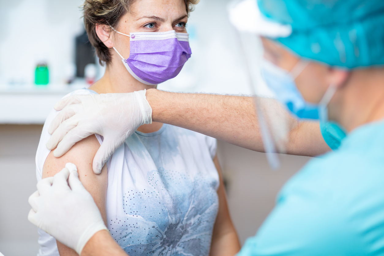 A young woman wearing a face mask sitting on a medical table as a doctor prepares to give her a vaccination.