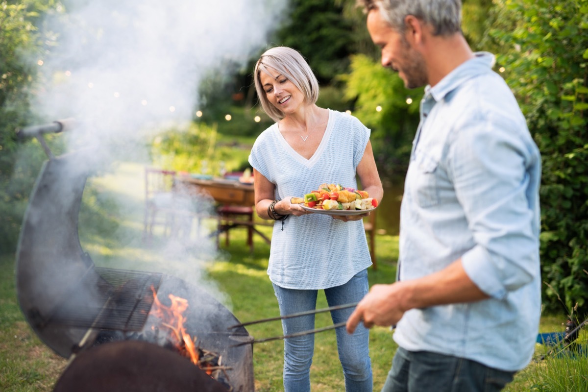 In summer. A nice couple prepares a bbq to welcome friends in the garden. She's holding a plate of grilled skewers