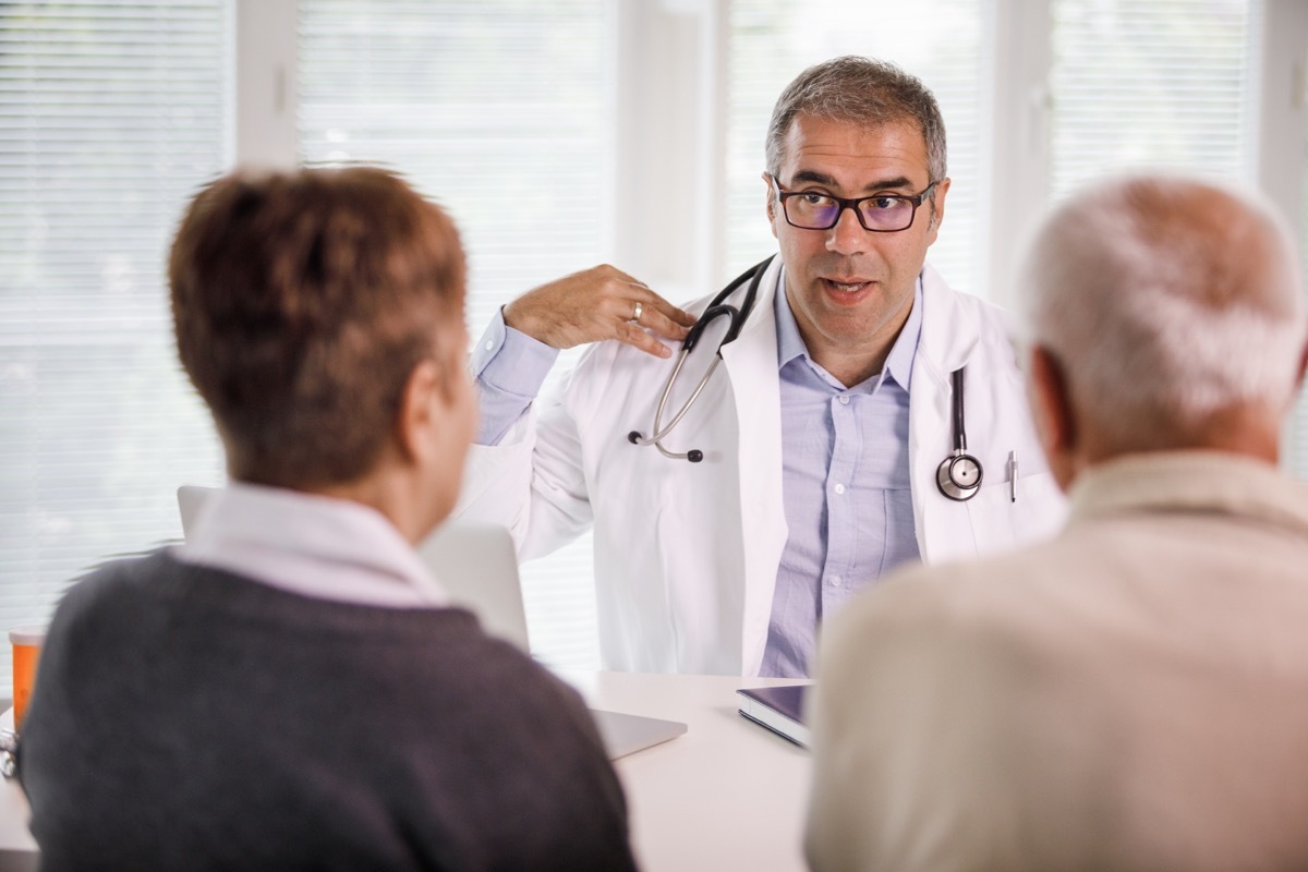 Selective focus of a mid-adult positive doctor explaining physical therapy exercises during medical consultation to an elderly couple sitting in front of him.