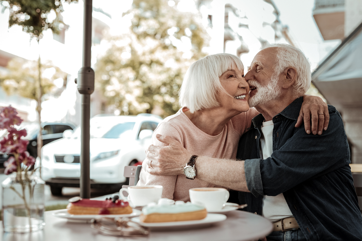 A happy senior couple kissing while sitting at an outdoor cafe with cake on the table