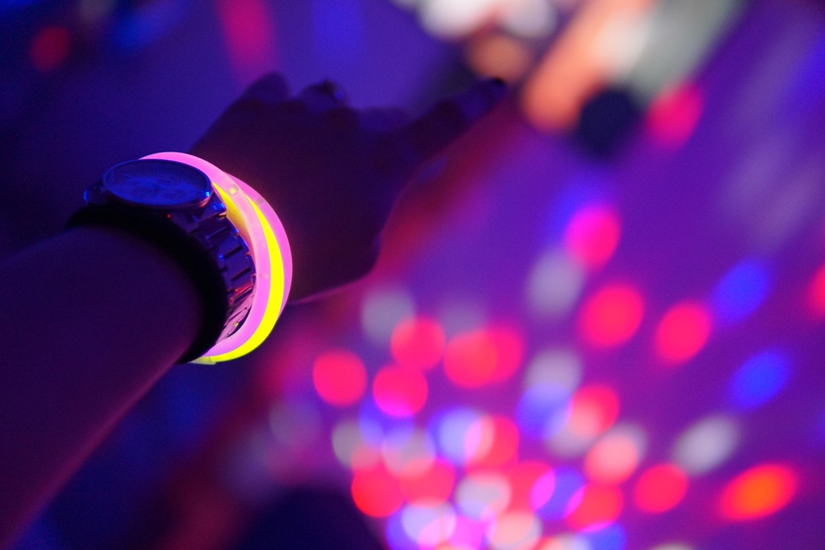 Hand with a watch and pink and yellow neon bracelets in the air during a neon party with blurry lights in the background - Image