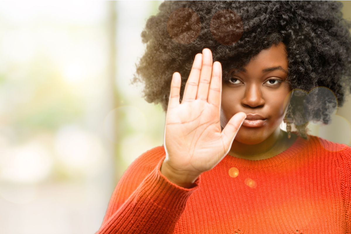 young Black woman wearing orange sweater holding up her hand to say 