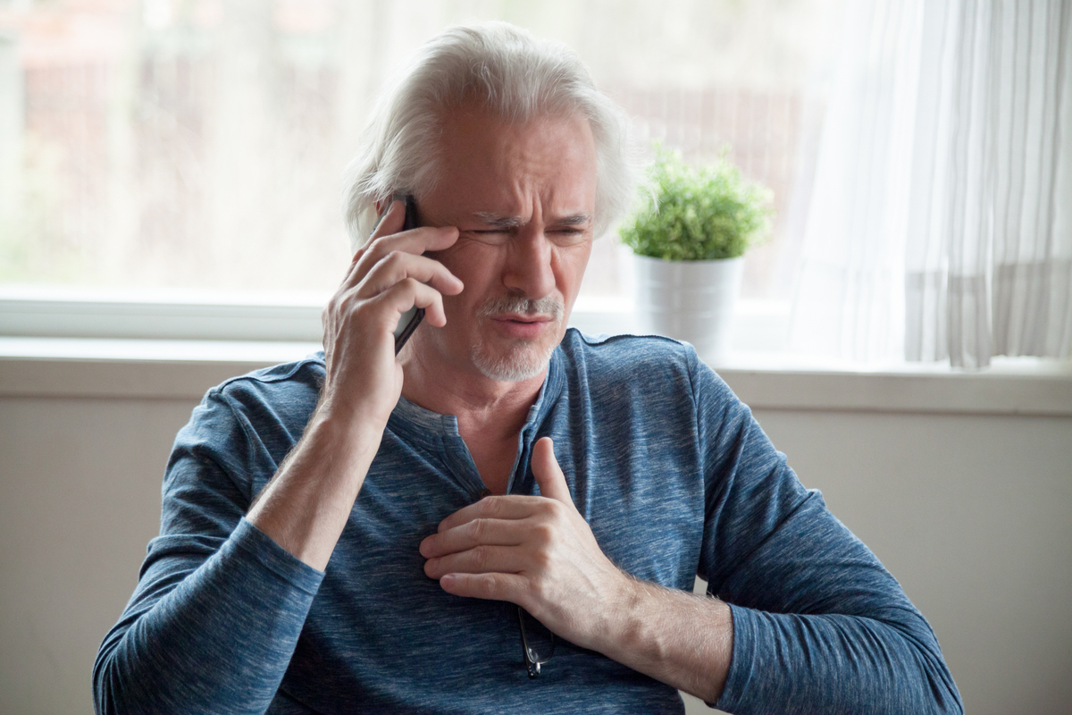 older white man with long hair clutching chest and talking on phone, looking worried