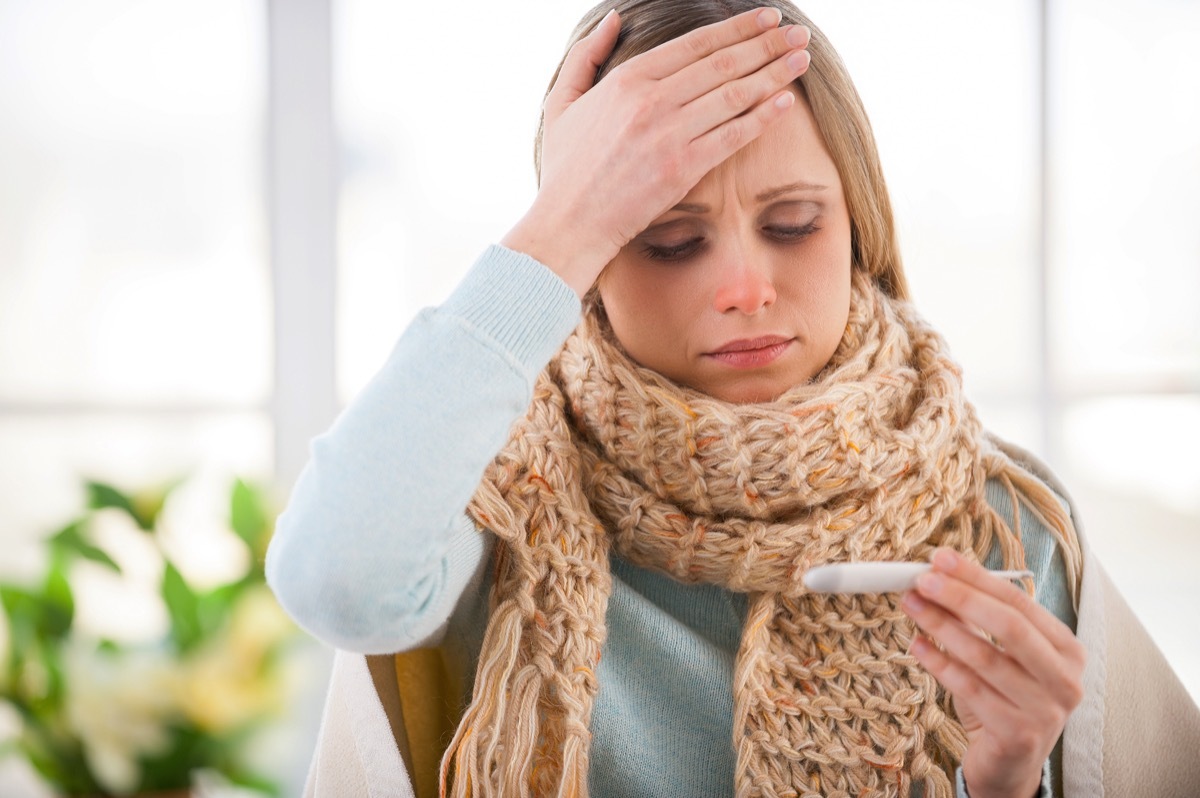 woman covered with plaid checking her body temperature while sitting in bed at her apartment