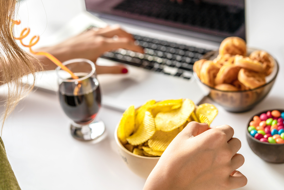 woman works at a computer and eats unhealthy food: chips, crackers, candy, waffles, soda