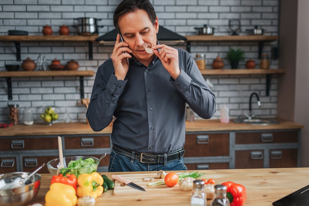 Adult man stand at kitchen table and talk on phone. He taste piece of mushroom. Man look down. Colorful vegetables lying on desk