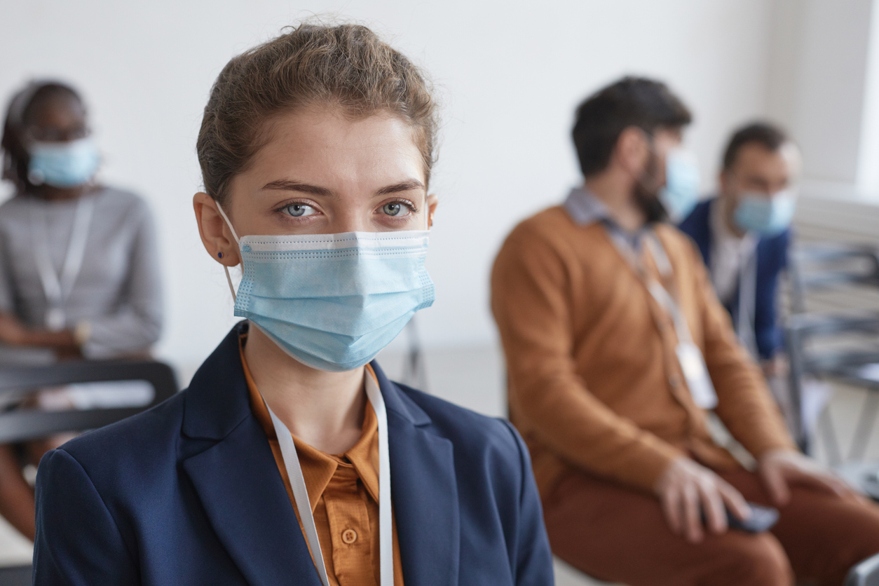 A young person wearing a suit and a face mask sits in front of other coworkers