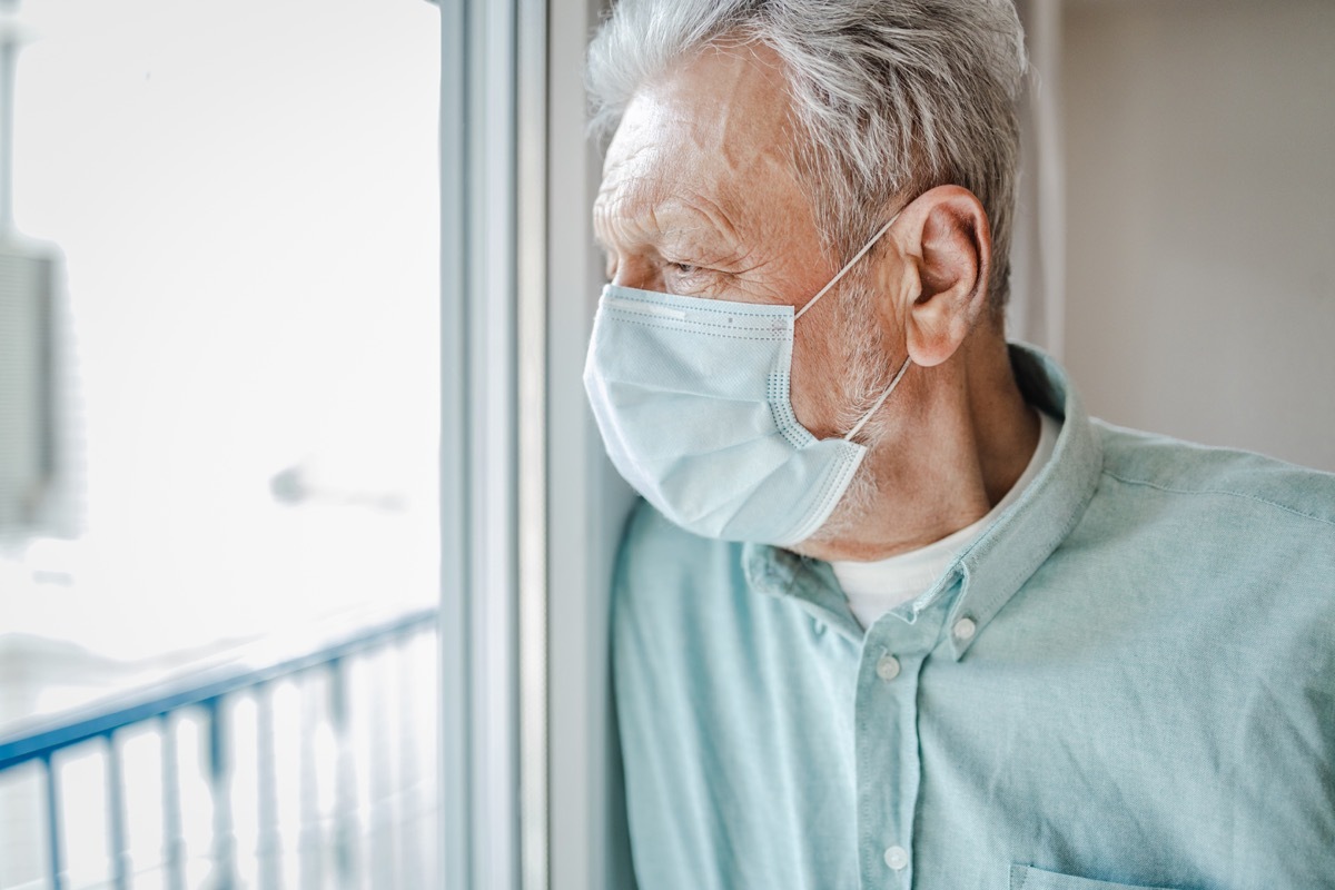 A senior man is at home by the window, he is wearing a protective face