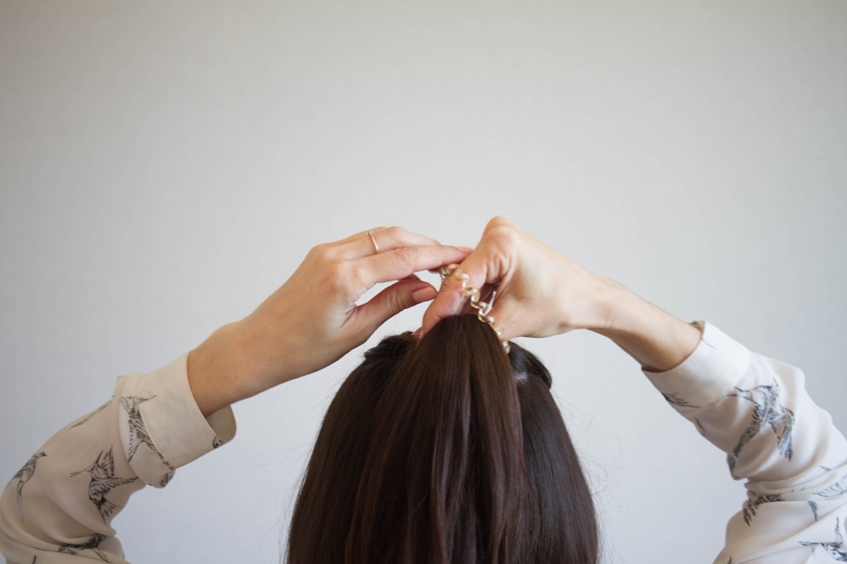 A young woman with brown hair ties her hair into a bun, viewed from the rear. Tutorial photo of simple hairstyle pinned half updo for long hair