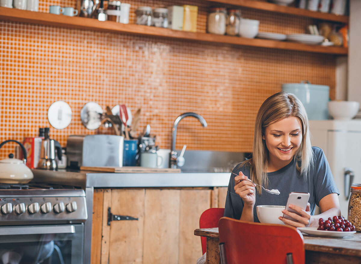 woman alone in kitchen looking at phone while eating