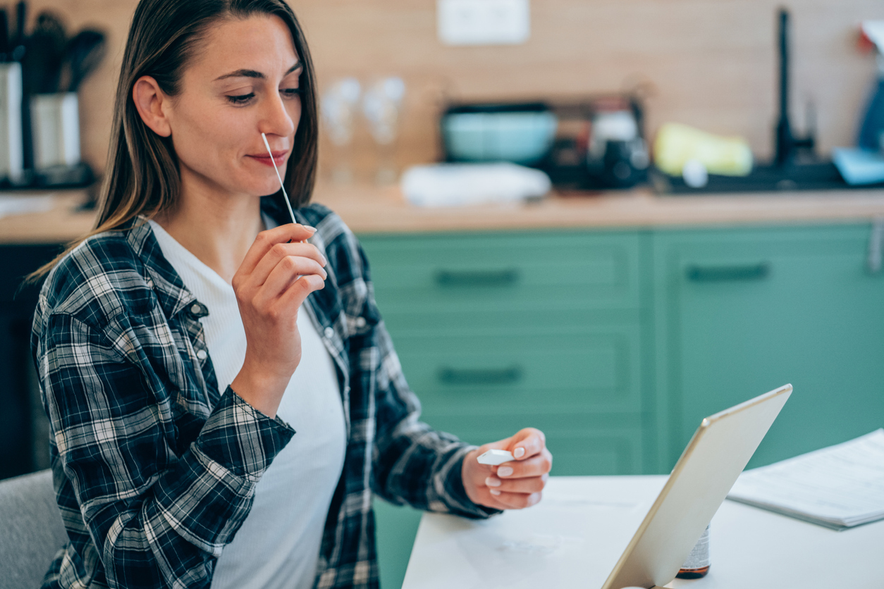 A young woman swabbing her nose for an at-home COVID test in front of her tablet