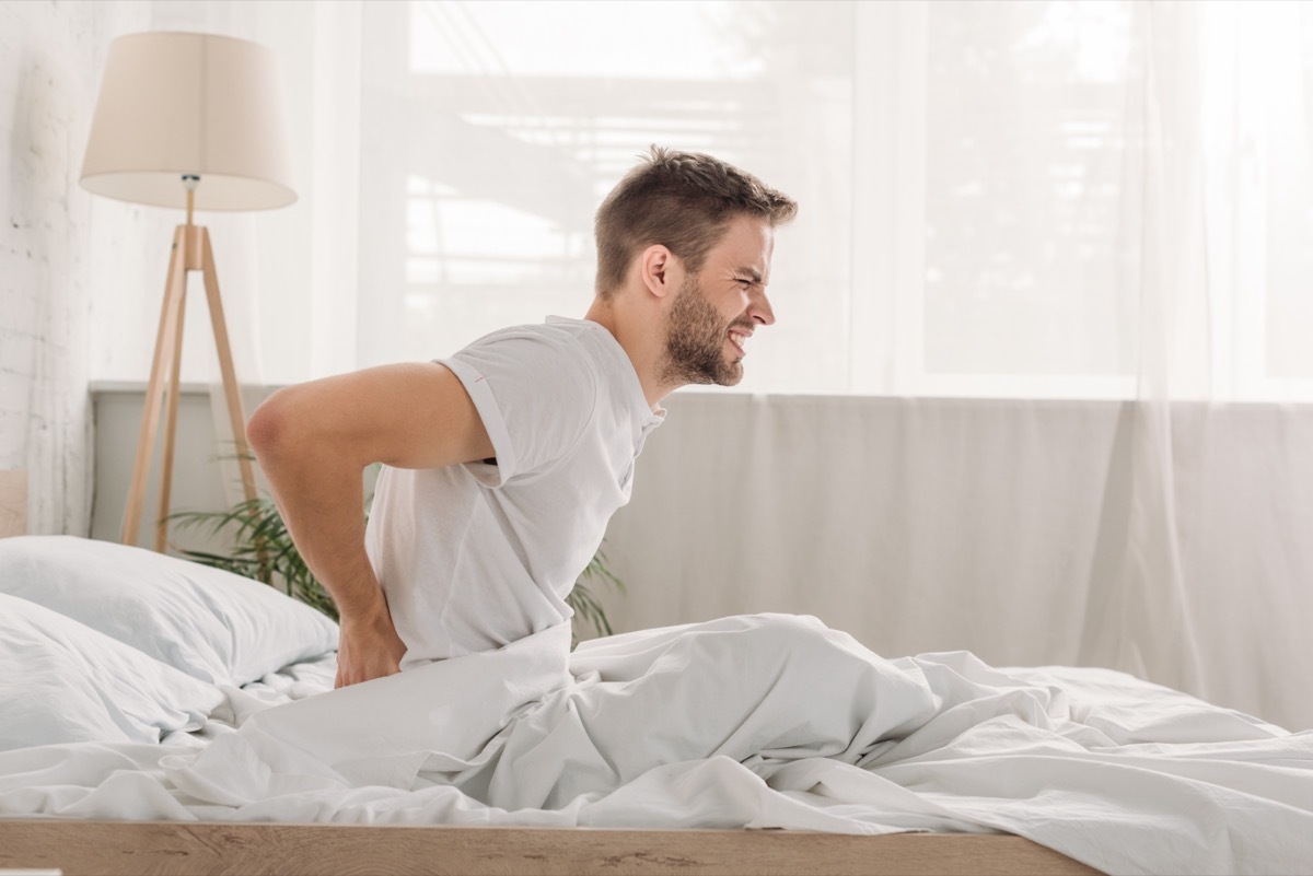 Side view of a frowned young man suffering from pain in loin while sitting on white bedding