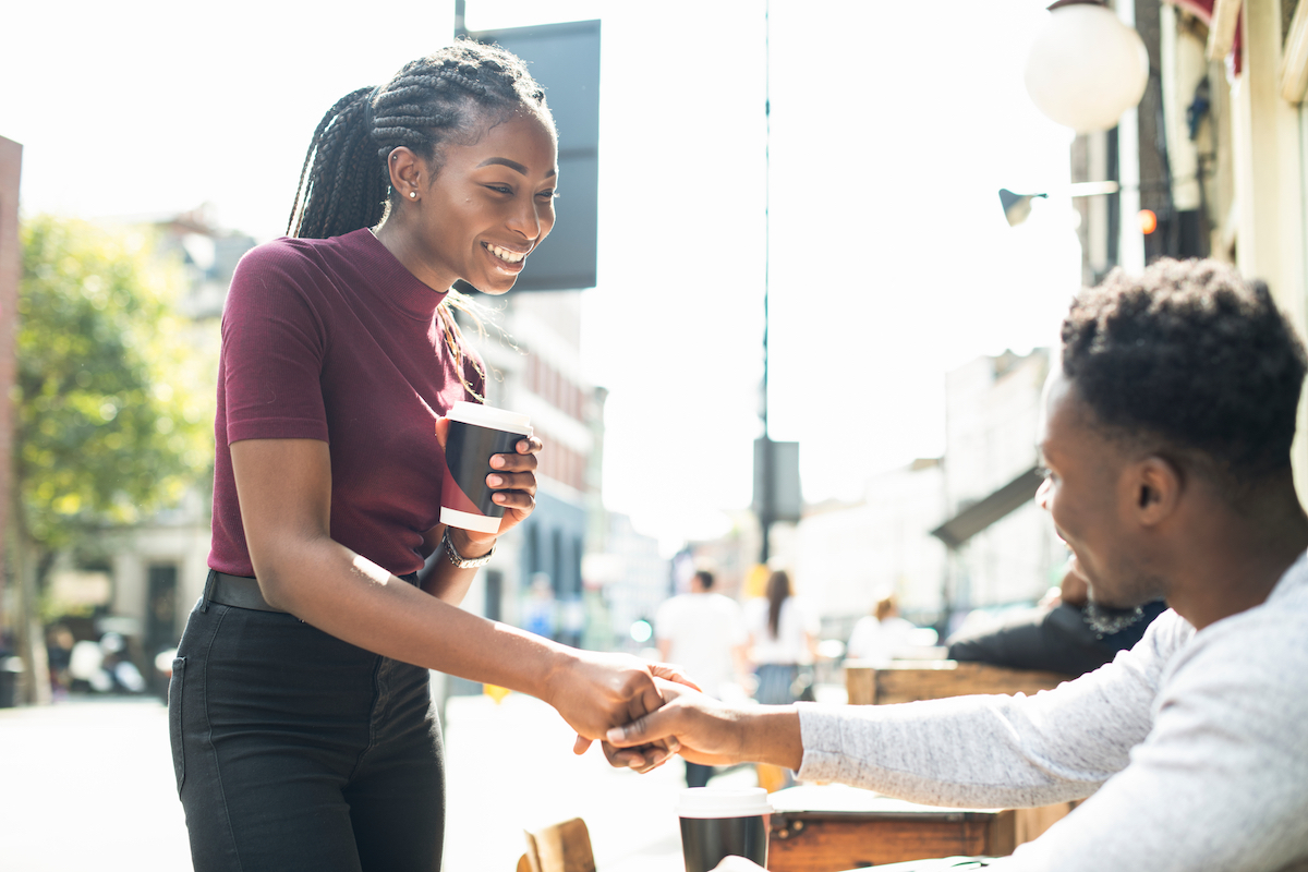 A man sitting at an outdoor restaurant table shakes hands with a woman who he's meeting for a first date.