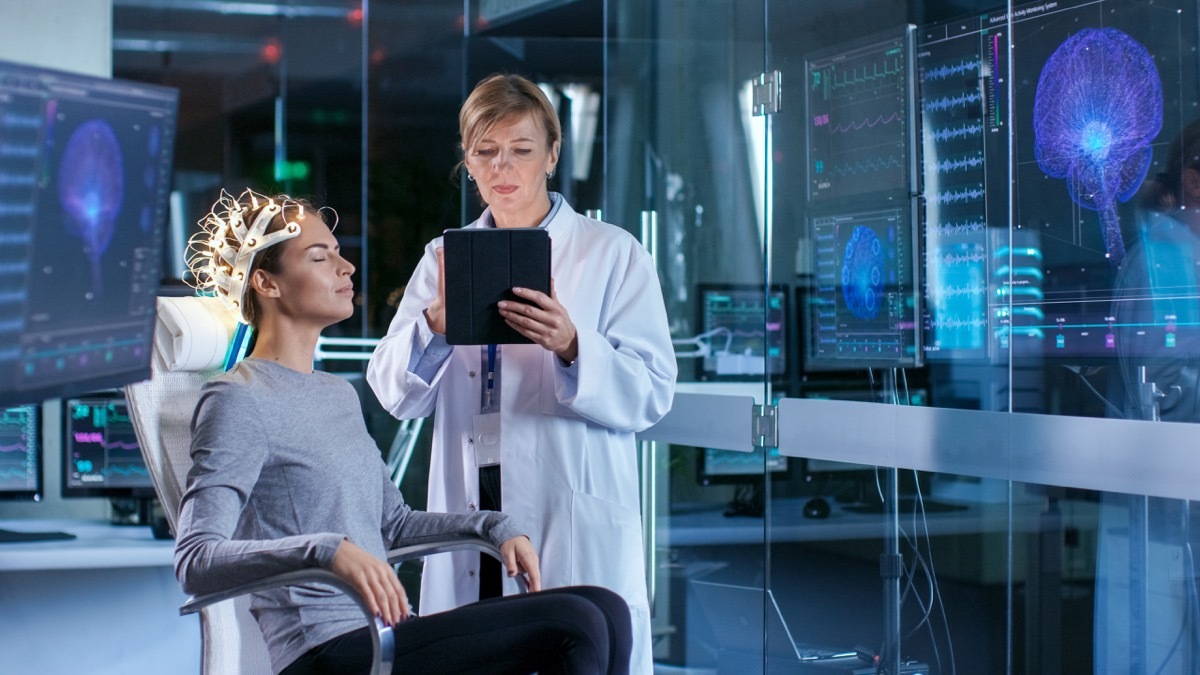 Woman Wearing Brainwave Scanning Headset Sits in a Chair while Scientist Adjusts the Device, Uses Tablet Computer. In the Modern Brain Study Laboratory Monitors Show EEG Reading and Brain Model. - Image