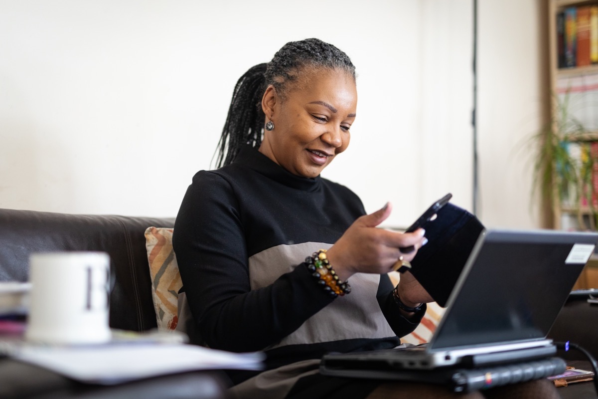 middle-aged woman wearing a black turtleneck with her hair in dreadlocks looking at phone and computer on couch