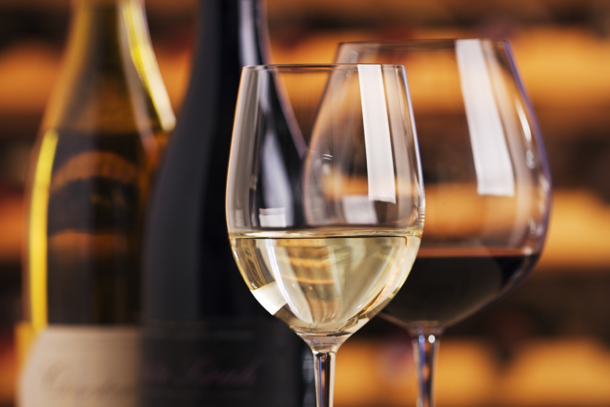 Wineglasses and wine bottles on display for serving a wine tasting. Red and white fermented grape beverages are poured into two sparkling glasses. Warm, orange brown wooden cellar wine racks are reflected and in soft focus in the background. The close-up view of the alcohol is indoors, with no people.