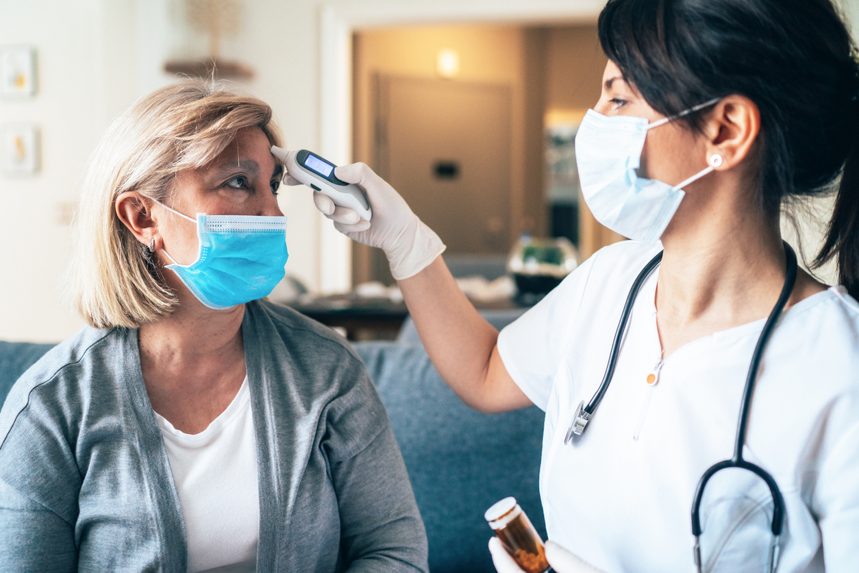 A female doctor takes a caucasian woman's temperature while both wear face masks.