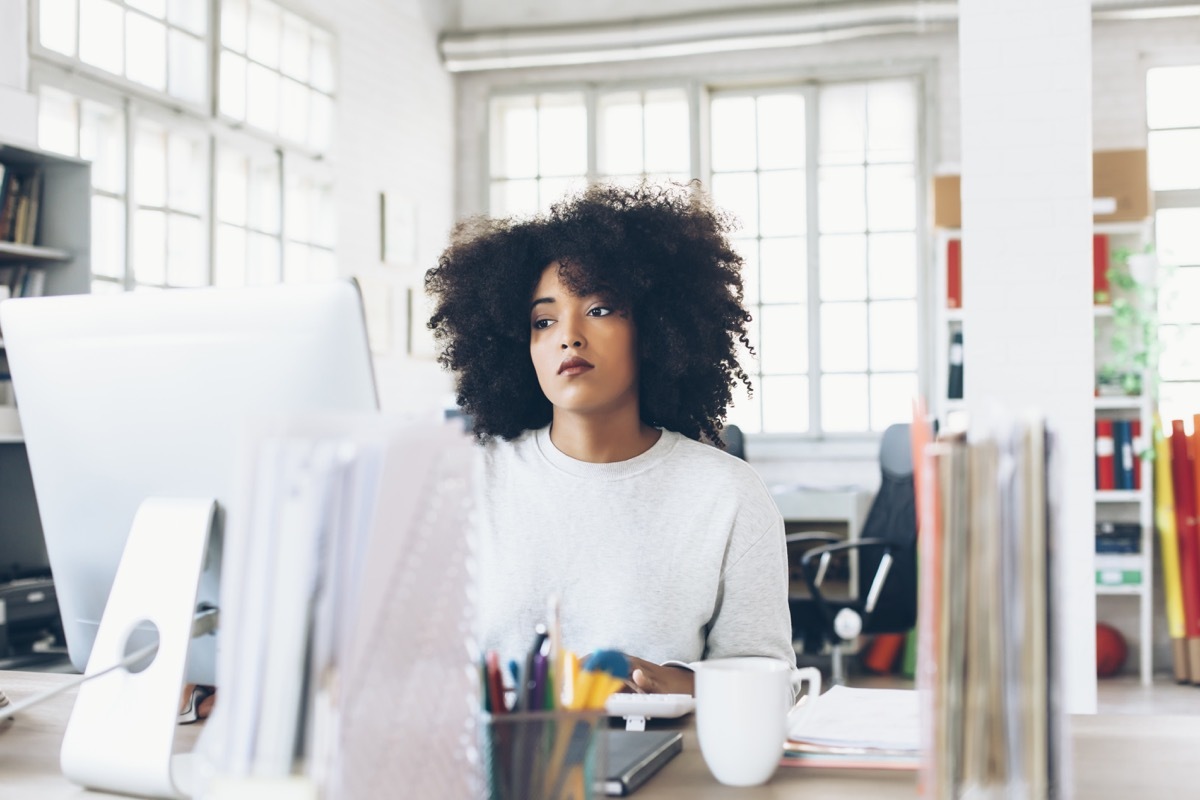 Tired young woman sitting on desk and using computer. Papers and tools, a cup on desk. Tall windows, shelves with folders, office seat on background.