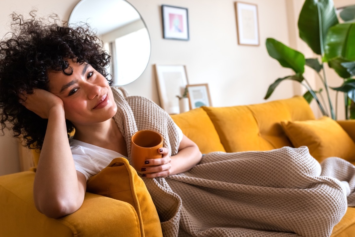 Happy African American woman relaxing at home lying on couch holding cup of warm coffee looking at camera. Copy space.