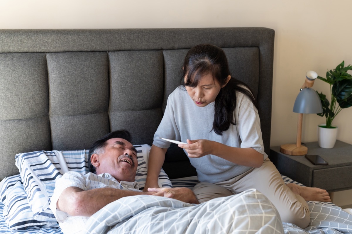 Senior man suffering from fever and stomach pain. The man pressing on stomach with painful expression. Wife checking body temperature of her ill husband on bed.