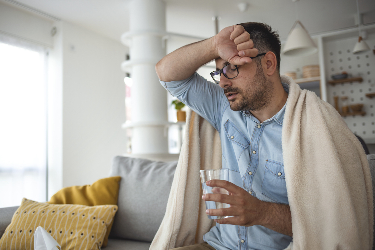 Young man suffering with fever and chills while sitting wrapped in a blanket on the sofa at home