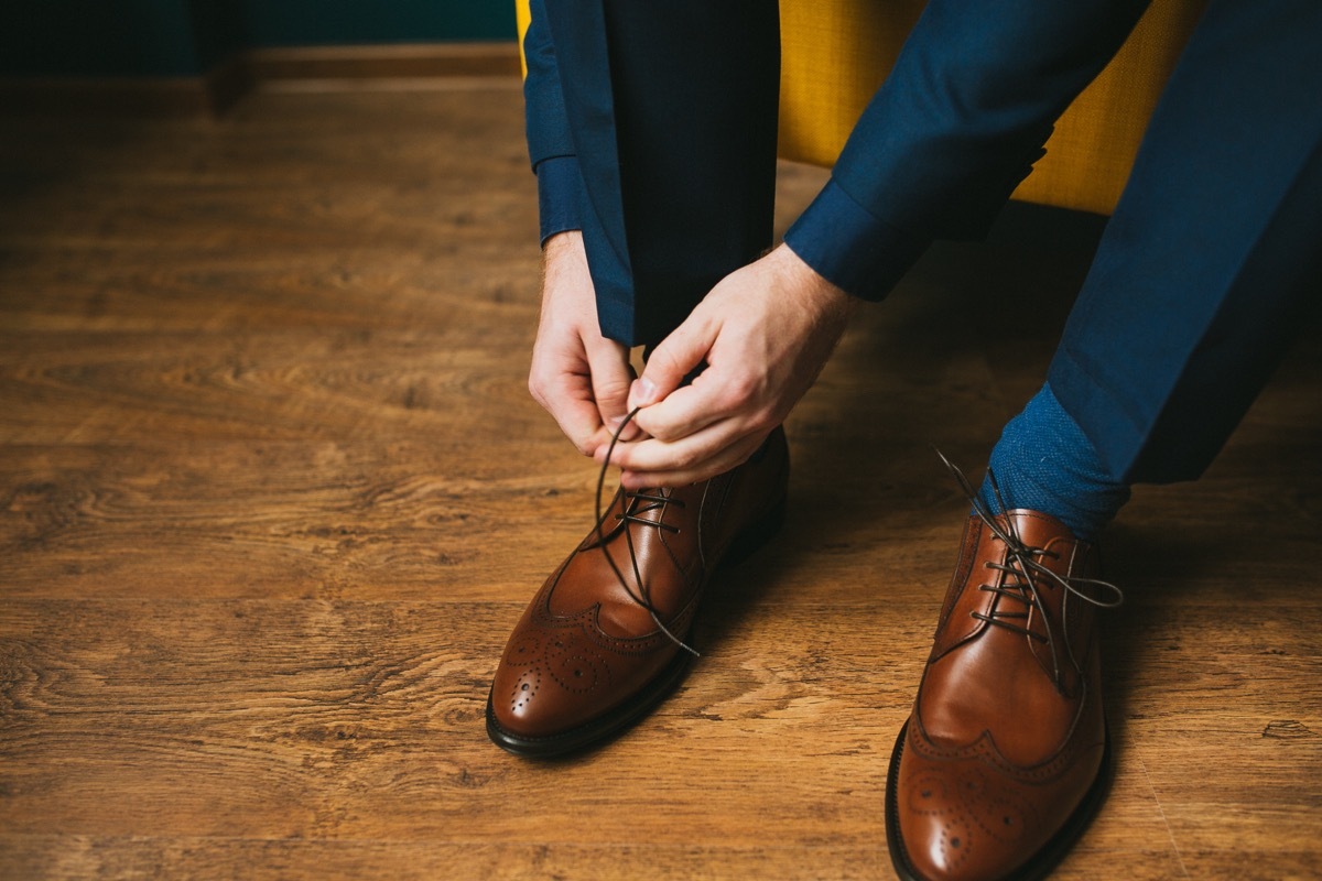 A man in a blue suit ties up shoelaces on brown leather shoes brogues on a wooden parquet background