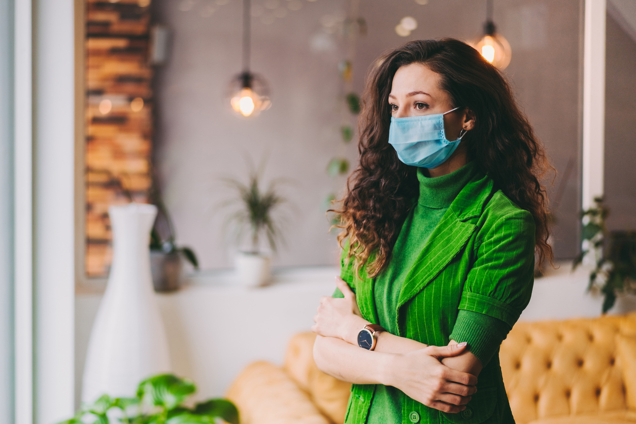 A young woman wearing a face mask stands inside an office lobby with her arms crossed.