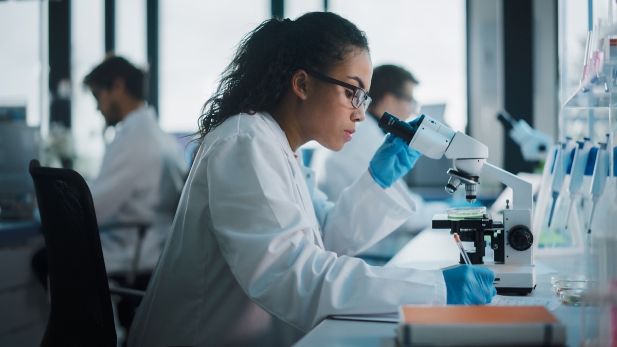 Medical Science Laboratory: Beautiful Black Scientist Looking Under Microscope Does Analysis of Test Sample. Diverse Team of Young Specialists, Using Advanced Technology Equipment.