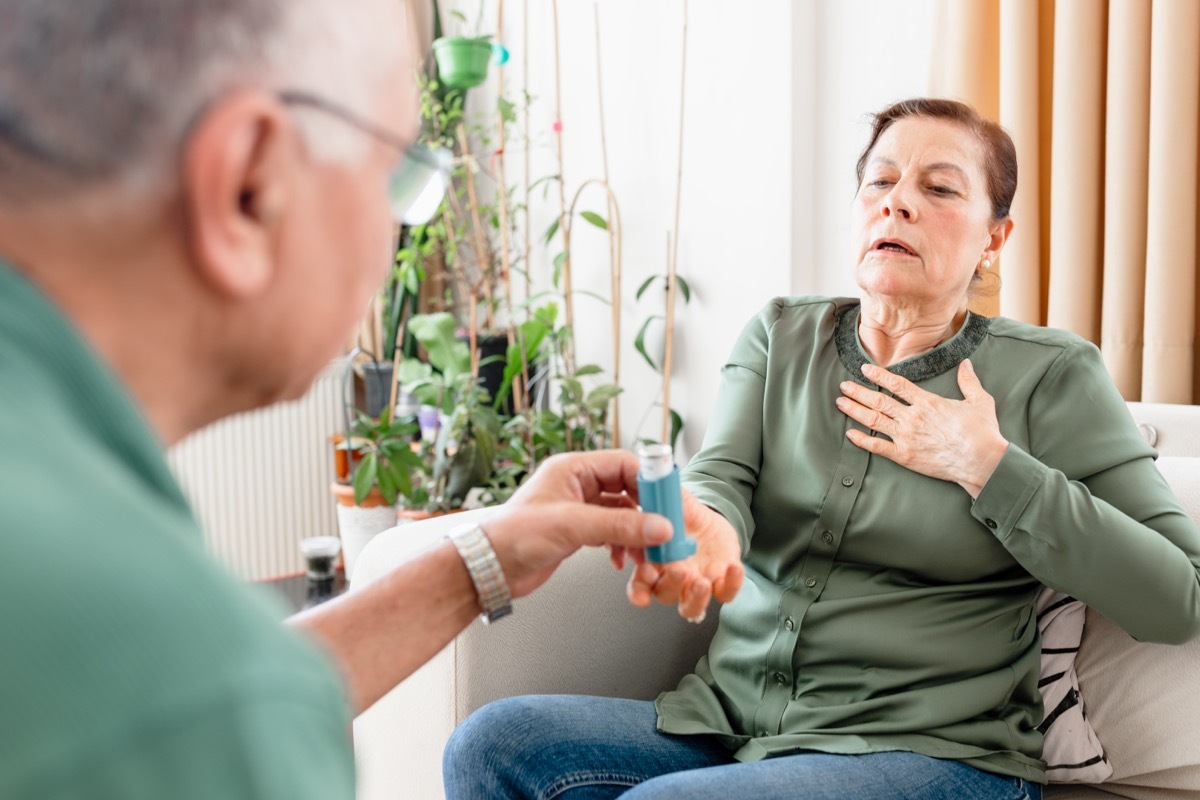 An older woman with asthma problems uses an asthma inhaler. she standing on sofa at home while her elderly husband helps her and gives him support to overcome the health problemsAn older woman with asthma problems uses an asthma inhaler. she standing on sofa at home while her elderly husband helps her and gives him support to overcome the health problems