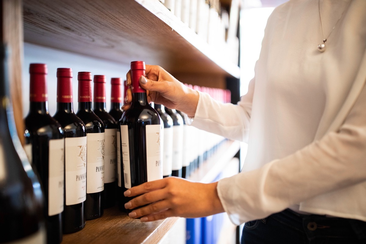 Close-up of female hands arranging wine bottles on shelf at wine shop. Woman employee at a wine store organizing the bottles on a stand.