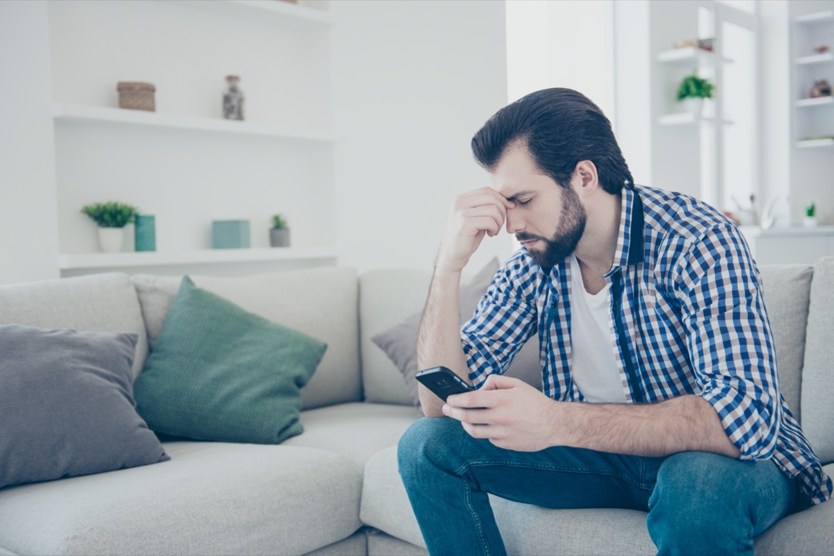 sad frustrated man with stubble in jeans, shirt holding fingers between close eyes using smart phone