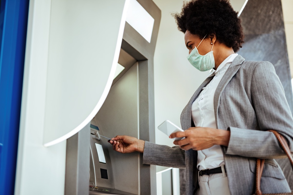 Low angle view of businesswoman inserting credit card and withdrawing cash at ATM while wearing protective mask on her face.